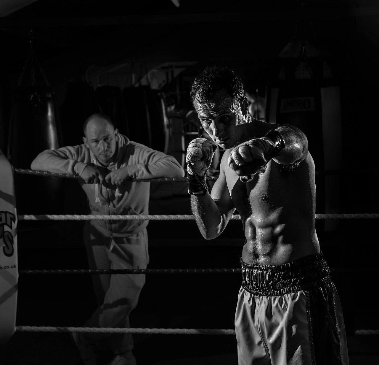 Boxeur torse nu en position de garde avec un coach observant en arrière-plan dans un ring de boxe.